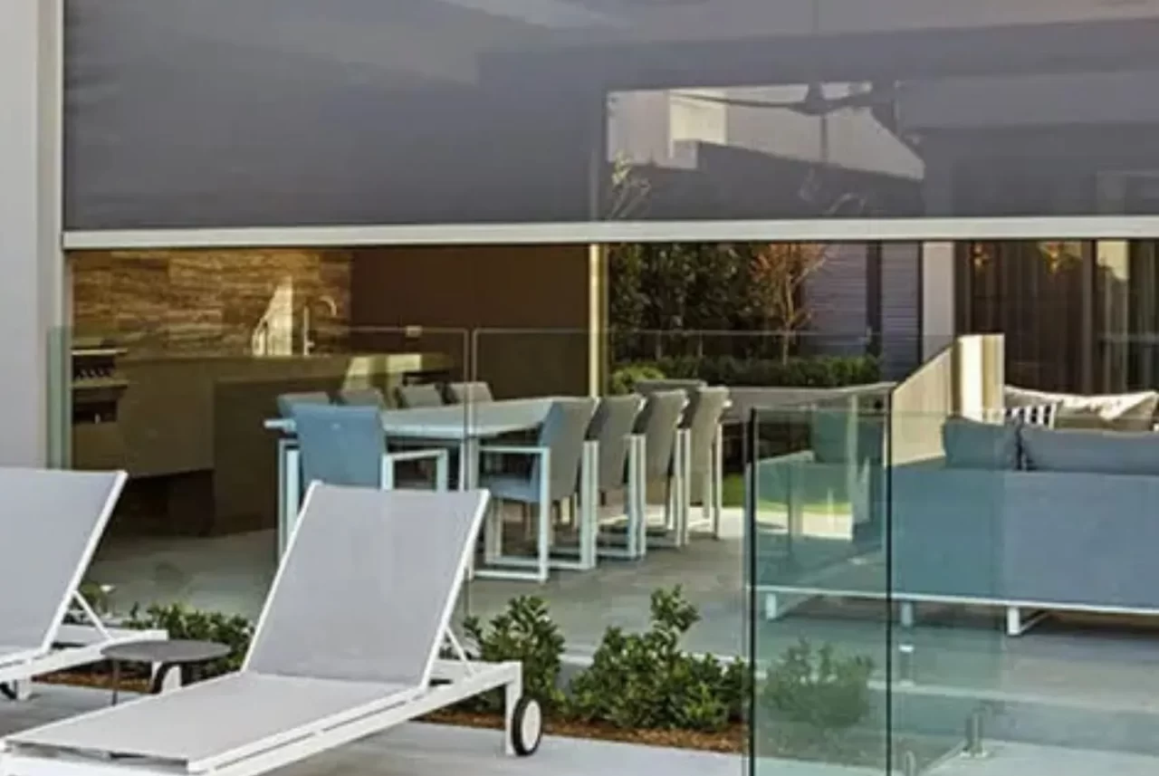 A Zipscreen blind raised above a glass-fenced poolside alfresco with a dining setting