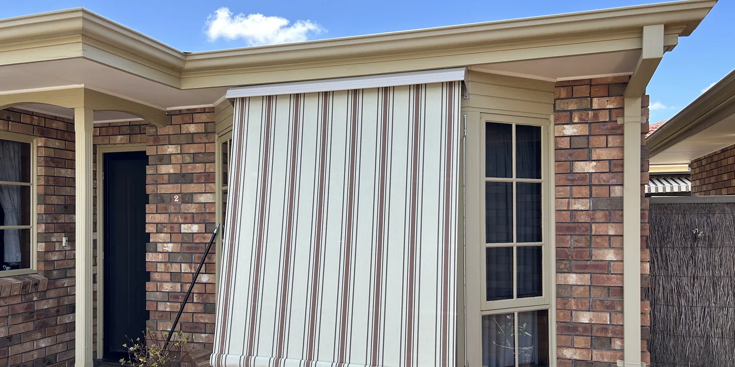 Two cream striped auto guide awnings lowered over the front windows of a brick home