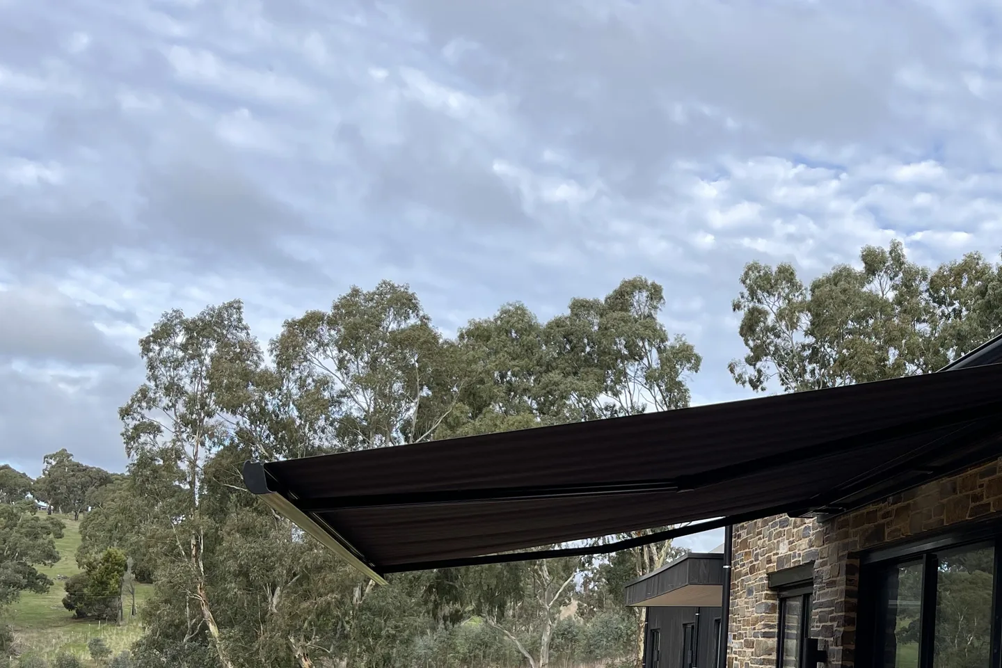 Dark folding arm awning extended along a stone-walled home facing a hillside lawn
