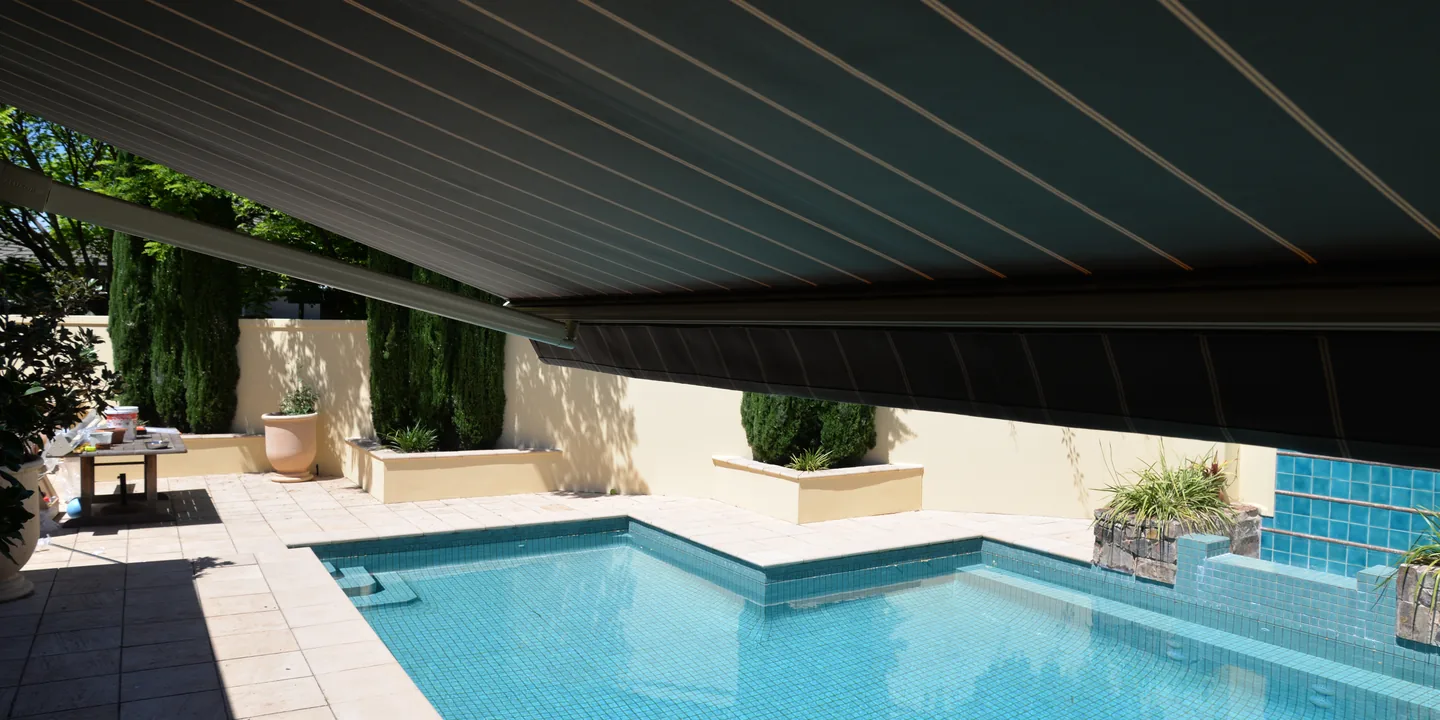 Charcoal striped folding arm awning extended over paving beside a backyard pool