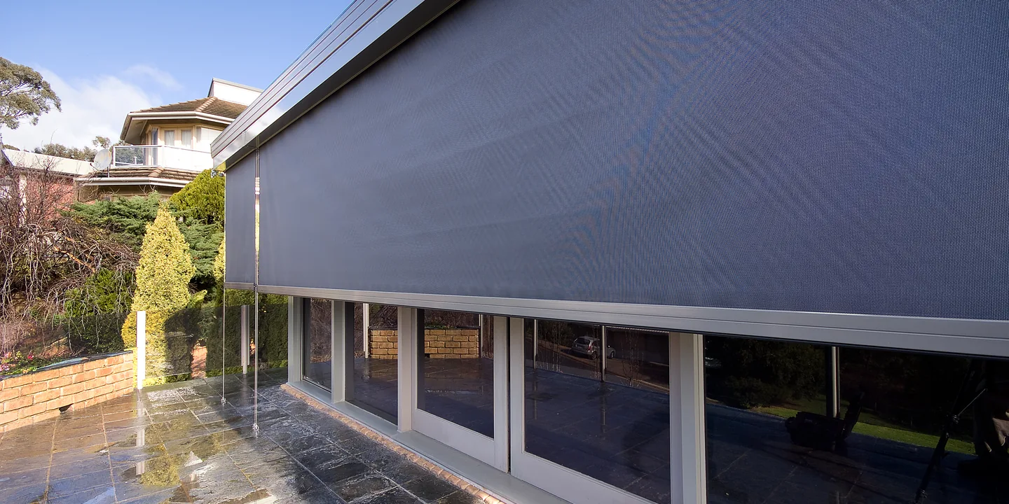 Charcoal wire guided outdoor blind lowered along a wall of glass doors on a wet stone terrace