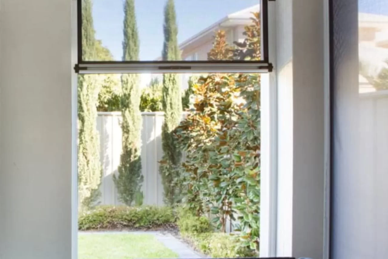 A dark Ziptrak blind raised to the head of a tall window, with the garden and trees outside