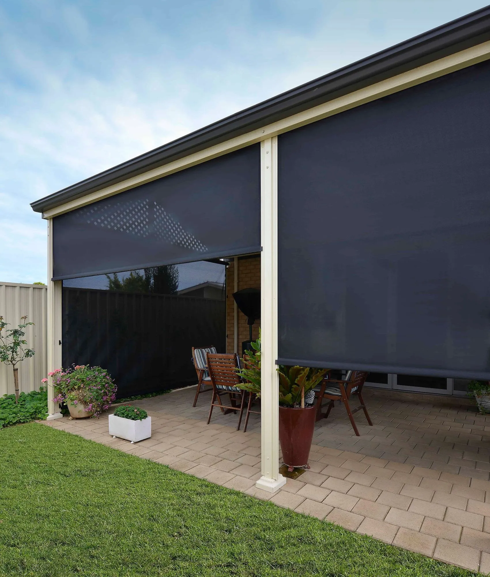 Two charcoal Ziptrak blinds between the posts of a paved patio, one lowered further than the other