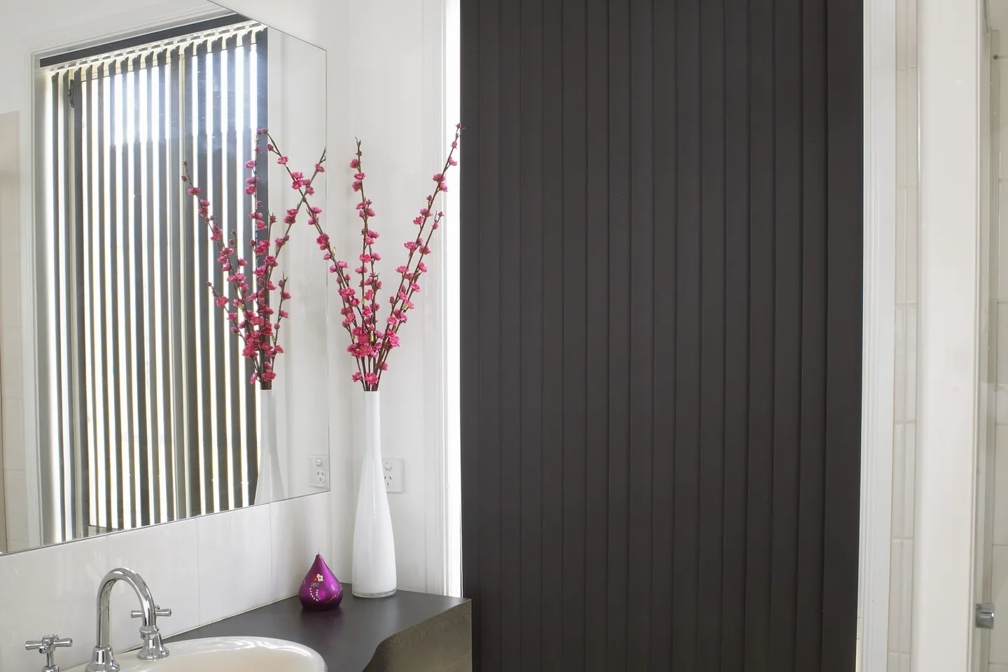 Dark vertical blind closed over a tall bathroom window beside the vanity