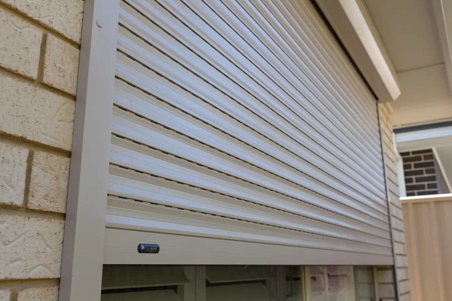 Close view of cream aluminium roller shutter slats and the side guide against a brick wall