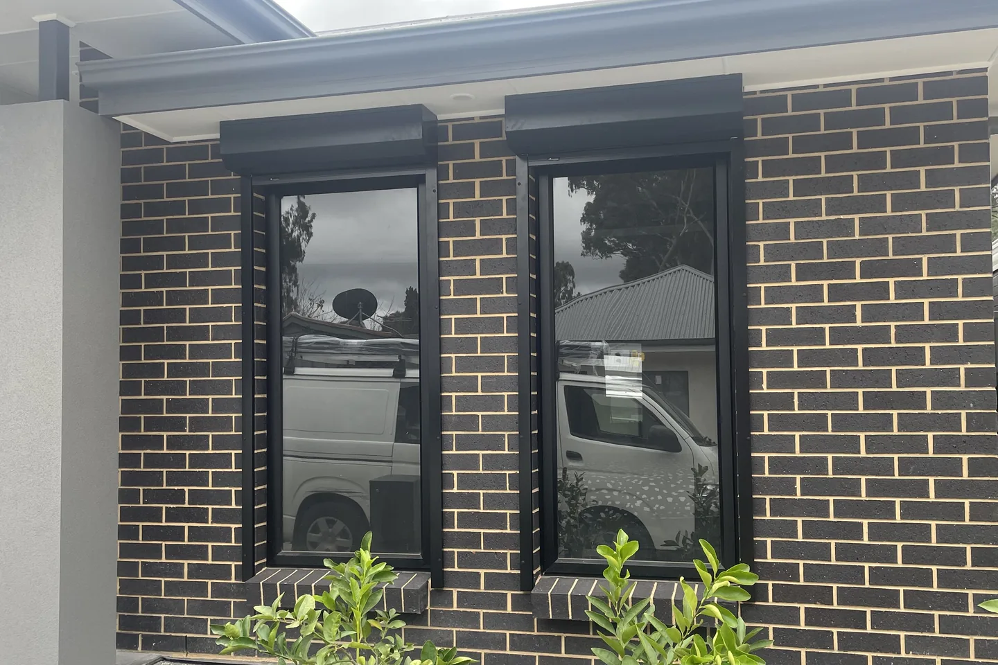 Two narrow windows with roller shutters raised into slim black headboxes on a dark brick wall