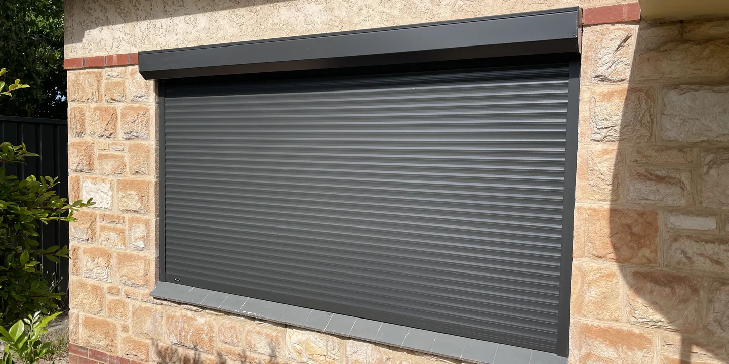 Charcoal aluminium roller shutter lowered over a wide window in a sandstone wall