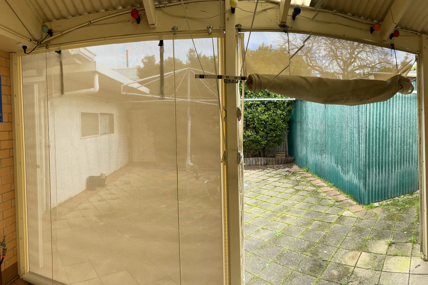 Pergola corner with a mesh outdoor blind lowered on one side and another rolled up and held above the opening