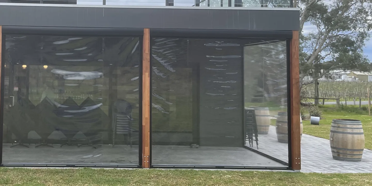 Clear zip roll up blinds lowered between timber posts on the ground floor of a pavilion beside a lawn