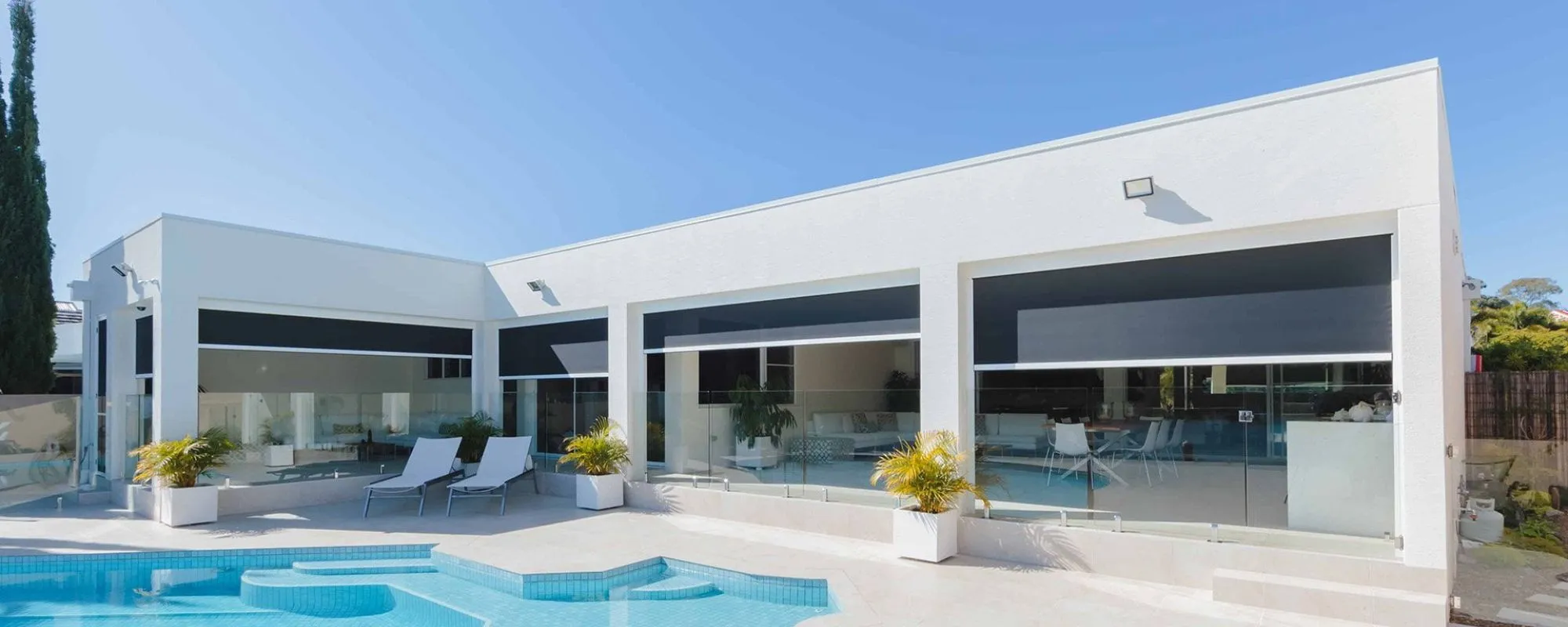 Zipscreen blinds across the alfresco openings of a white poolside home