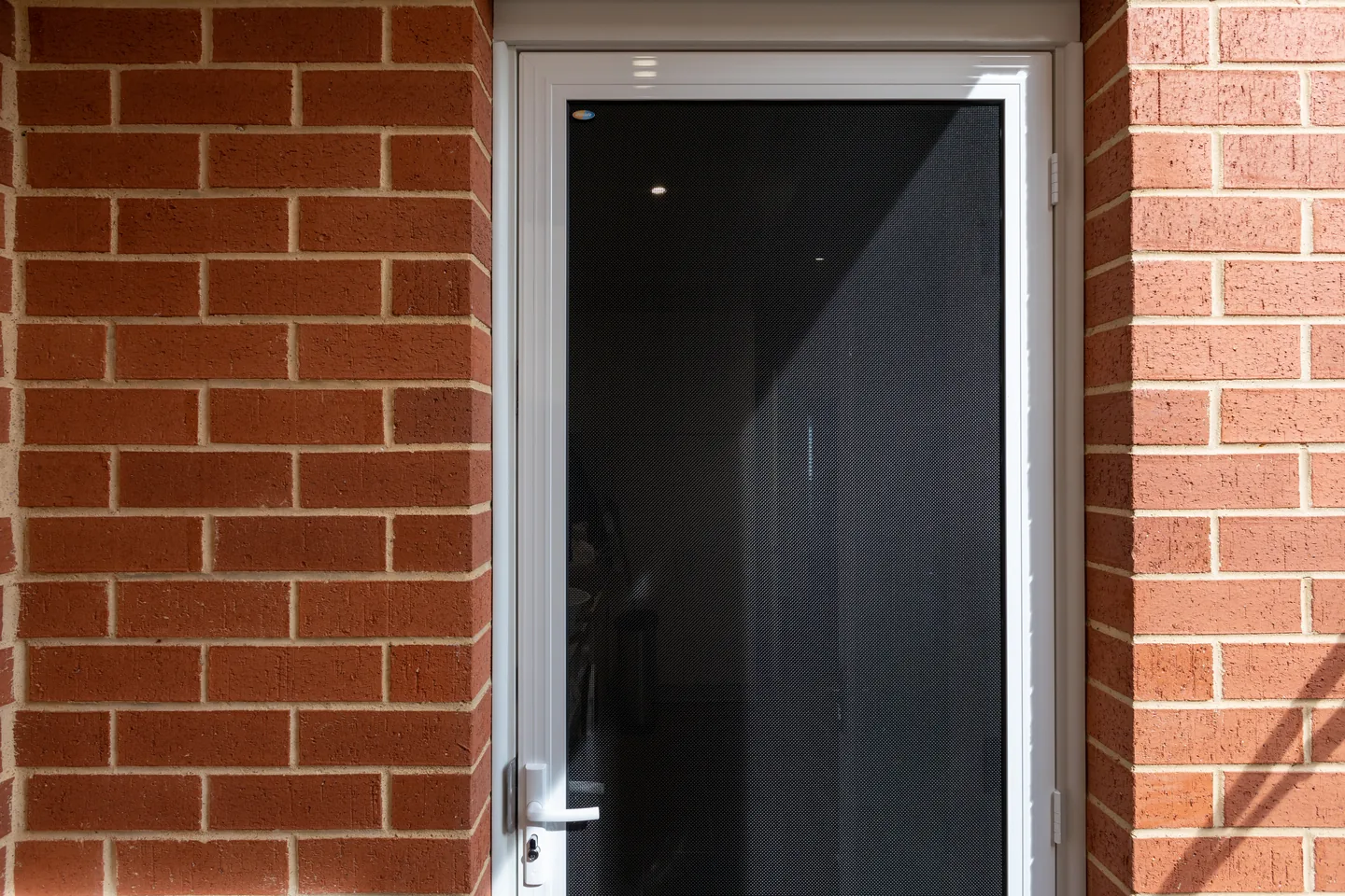 Crimsafe security door with a white frame set into a red brick entry reveal, lever handle and keyed lock at the side