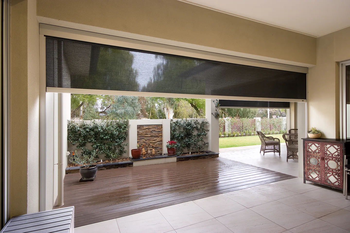 Covered timber deck looking out to a garden, with a mesh outdoor blind partly lowered across the opening