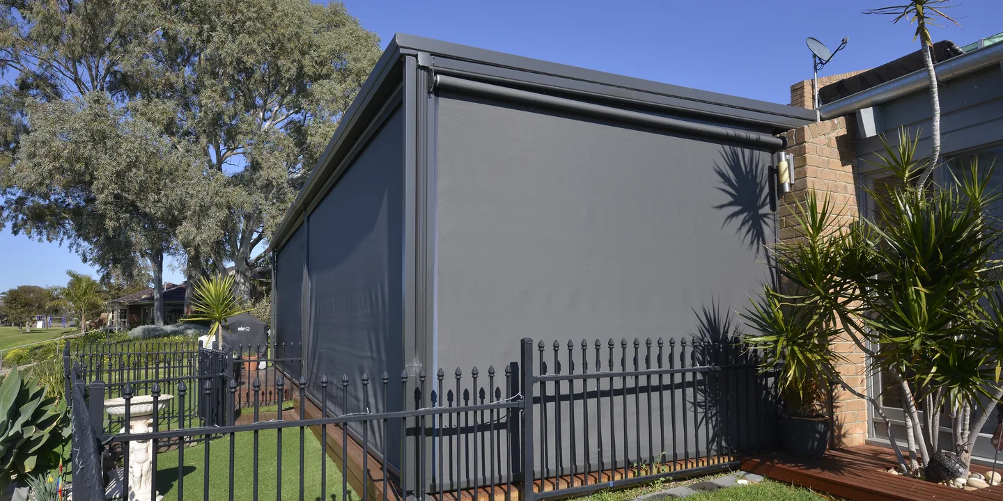 Dark mesh Ziptrak outdoor blinds lowered around a charcoal-framed outdoor room beside a lawn
